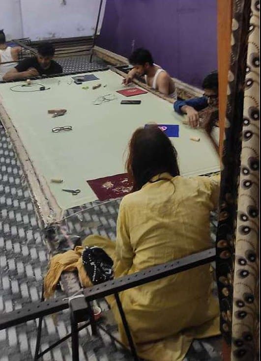 A woman seated at a large embroidery frame as embroiderers work across it.