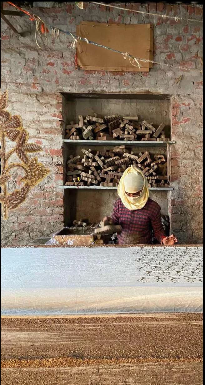A printer at a long table in a brick workshop, shelves of carved wooden blocks behind.