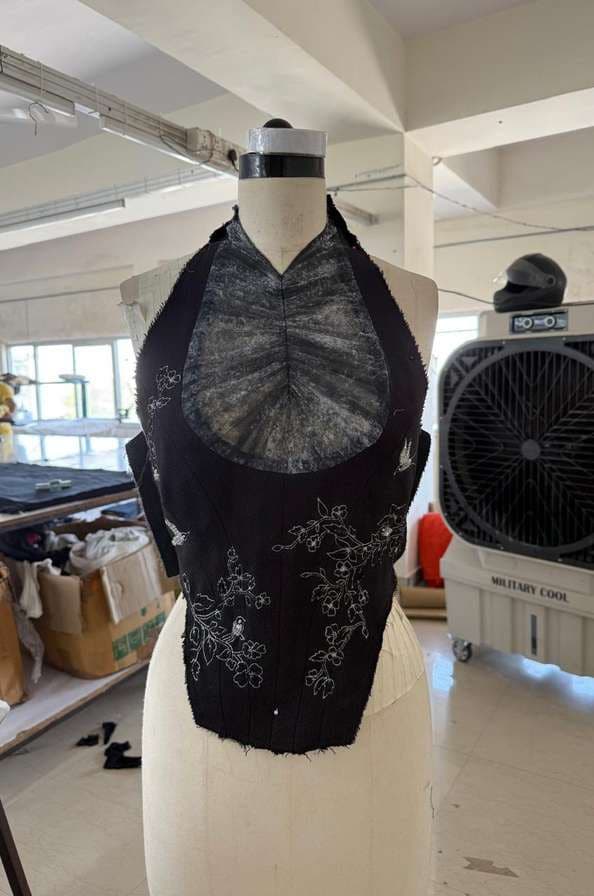 A black corset on a dress form, gathered sheer front panel, embroidered with white zari birds and stems.